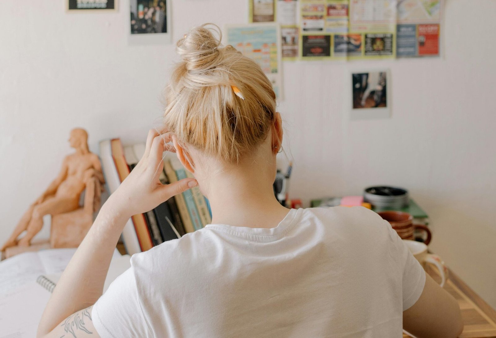 Blonde artist with a messy bun sits at a cluttered desk, appearing frustrated or lost in thought; a real moment of stalled art motivation and inspiration.